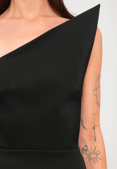Nærbillede af en person iført en sort asymmetrisk kjole med synlige minimalistiske tatoveringer på overarmen og skulderen.