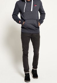 Sudadera gris con cordones blancos, bolsillo frontal y pequeño logo rojo; combinada con pantalones ajustados negros y zapatos negros.