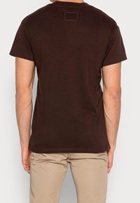 Braunes Baumwoll-T-Shirt mit kurzen Ärmeln, lockerer Passform und einem kleinen quadratischen Etikett, das auf der Rückseite des Halsbereichs angenäht ist.