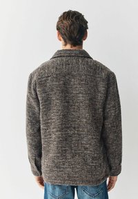 Chaqueta marrón texturizada con cuello, que presenta un patrón tejido. Las mangas son largas y se acompaña de unos vaqueros de mezclilla azules.