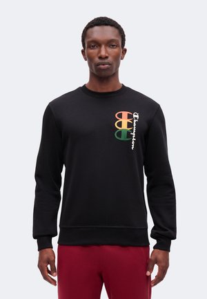 Mann trägt schwarzen langärmeligen Champion-Sweatshirt mit mehrfarbigem Logo auf der Brust und weinrote Hose, steht vor einfachem Hintergrund.
