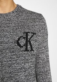 Gros plan d'une personne portant un pull en maille noir et blanc avec un col rond et les initiales noires "CK" sur la poitrine.