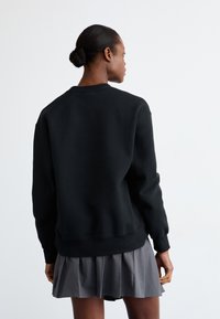 Sudadera negra de manga larga con puños acanalados. Presenta un cuello redondo y un corte holgado, combinado con una falda gris plisada.