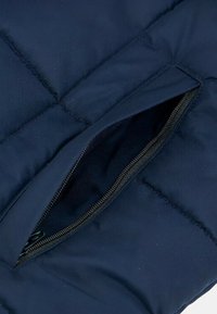 Giacca di jeans blu con una sezione che presenta una tasca con cerniera aperta. Il materiale ha una consistenza liscia con cuciture visibili e una cerniera scura.