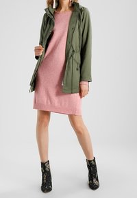 Veste légère vert olive avec taille à cordon sur une robe en tricot rose. Bottines noires ornées d'un motif floral.