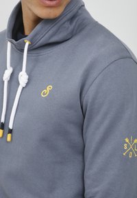 Felpa grigia con colletto alto, lacci bianchi e dettagli gialli. Presenta un piccolo logo ricamato sul petto e un dettaglio con motivo sulla manica.
