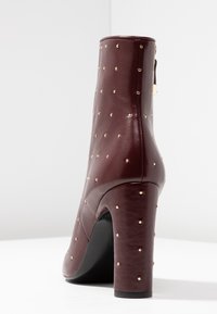 Tmavě burgundy kožené kotníkové boty se zlatými hřebíky, blokovým podpatkem a zadním zipem, vyfocené na bílém pozadí.