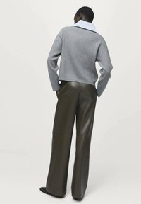 Persona que lleva una camisa de cuello azul claro debajo de un suéter gris y pantalones anchos oscuros, de pie con las manos en los bolsillos, mirando hacia el lado opuesto.