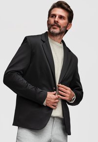 Homme avec barbe ajustant un blazer noir par-dessus un pull clair et un pantalon gris, regardant sur le côté devant un fond uni.