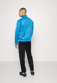 Modrá ľahká bunda s vysokým golierom, dlhými rukávmi a elastickými manžetami, v kombinácii s čiernymi športovými nohavicami a čiernymi topánkami.