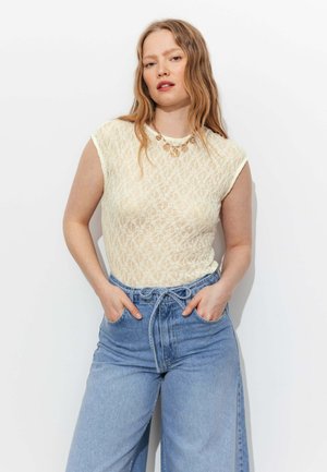 Femme aux longs cheveux ondulés portant un top sans manches en dentelle crème, un collier en or et un jean taille haute bleu clair à jambes larges, les mains dans les poches.