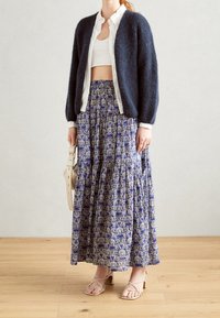 Námořnický chlupatý cardigan, bílá košile s knoflíky, crop top a dlouhá modrá vzorovaná sukně s patry. Světle barevné sandály na řemínky a béžová taška.