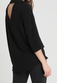 Svart blus med krage, trekvartsärmar och detalj med öppning i ryggen. Slät textur, avslappnad passform och rundad nederkant.