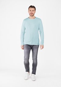 Jasnoniebieska koszula z długim rękawem i okrągłym dekoltem, w zestawieniu z szarymi zwężanymi dżinsami w stylu distressed oraz białymi trampkami, na jednolitym tle.