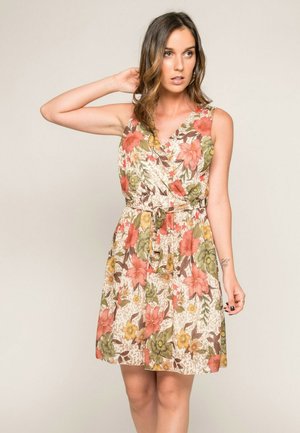 Femme portant une robe sans manches à fleurs roses, jaunes et vertes, posant avec une main derrière la tête sur un fond beige.