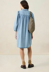 Femme portant une robe en denim bleu clair, des chaussettes marron, des mocassins sombres, et portant un sac à bandoulière à imprimé léopard, se tenant devant un fond uni.