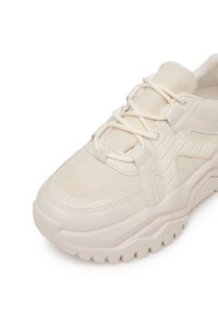 Zapatilla de color beige claro con parte superior de tela texturizada, suela de goma gruesa y elementos de diseño en capas con costuras visibles y acentos.