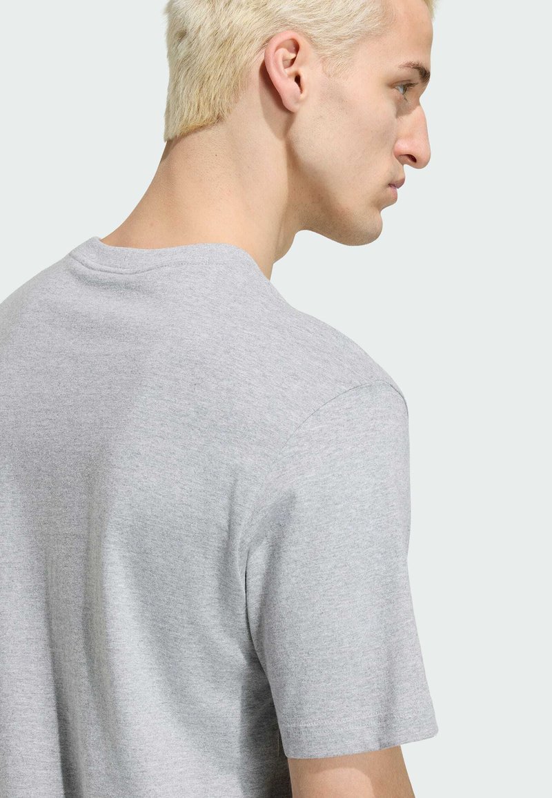 Ung person med kort bleget hår iført en ensfarvet lysegrå T-shirt, vist fra ryggen og siden mod en neutral baggrund.