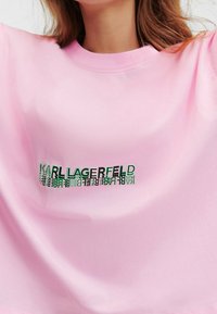 Žena má na sobě světle růžové tričko s logem "KARL LAGERFELD" ve zeleném a černém textu umístěným na hrudi.