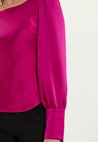 Blouse en soie fuchsia avec des manches longues et bouffantes, un col rond et des détails froncés aux poignets, accompagnée de boutons noirs en dessous.