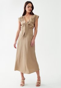 Robe beige, longueur genou avec de courtes manches flottantes et un détail de taille nouée. Tissu lisse et fluide. Portée avec des talons à lanières assortis.