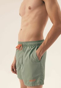 Zielone kąpielówki z elastycznym pasem w talii i pomarańczowym sznurkiem. Posiadają boczne kieszenie i małe logo na dolnej części nogawki.