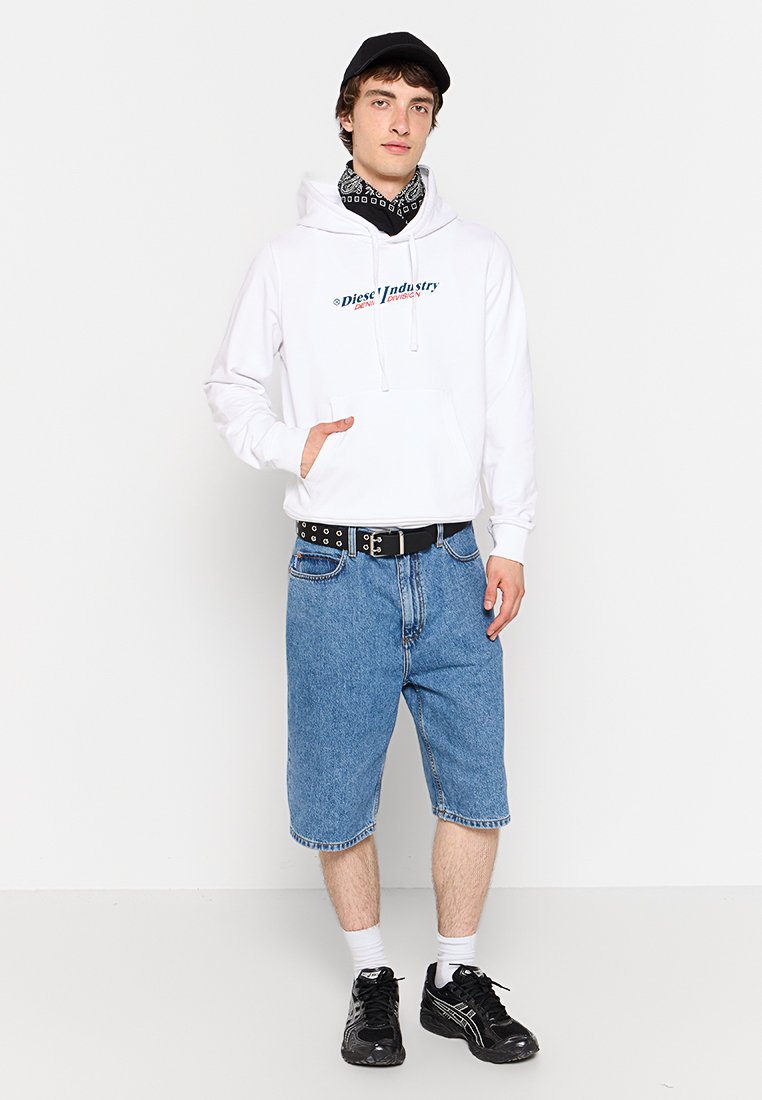 Giovane uomo che indossa un hoodie bianco Diesel, un cappello nero, un bandana nero, pantaloni shorts in denim, una cintura nera, calzini bianchi e sneakers nere, in piedi contro un muro bianco.
