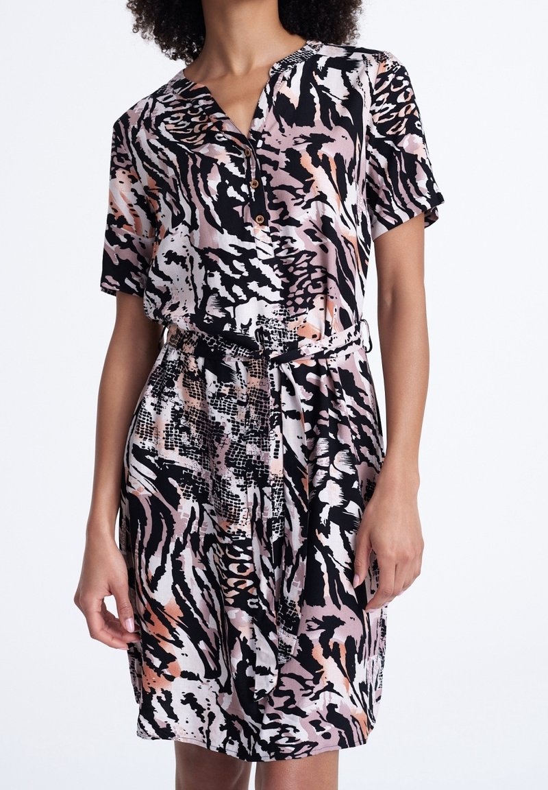 Femme portant une robe ceinturée à manches courtes avec un motif abstrait d'imprimé animal noir, rose et blanc, debout devant un fond uni.