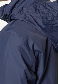 Chaqueta impermeable en color marino con textura suave y brillante; cuenta con un cuello alto, detalles cosidos y cordón ajustable en la capucha.