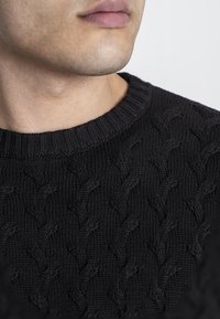 Suéter negro de punto con un patrón de trenzas y un cuello redondo acanalado, que presenta un acabado texturizado. Primer plano de la parte superior de la prenda.