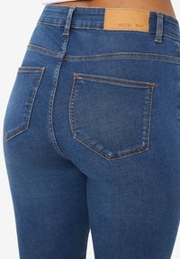 Gros plan sur une personne portant un jean denim bleu foncé avec des poches arrière et une étiquette en cuir de la marque "Noisy May" sur la ceinture.