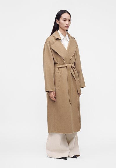 Femme portant un long manteau camel ceinturé, une chemise blanche et un pantalon large beige, debout sur un fond uni.