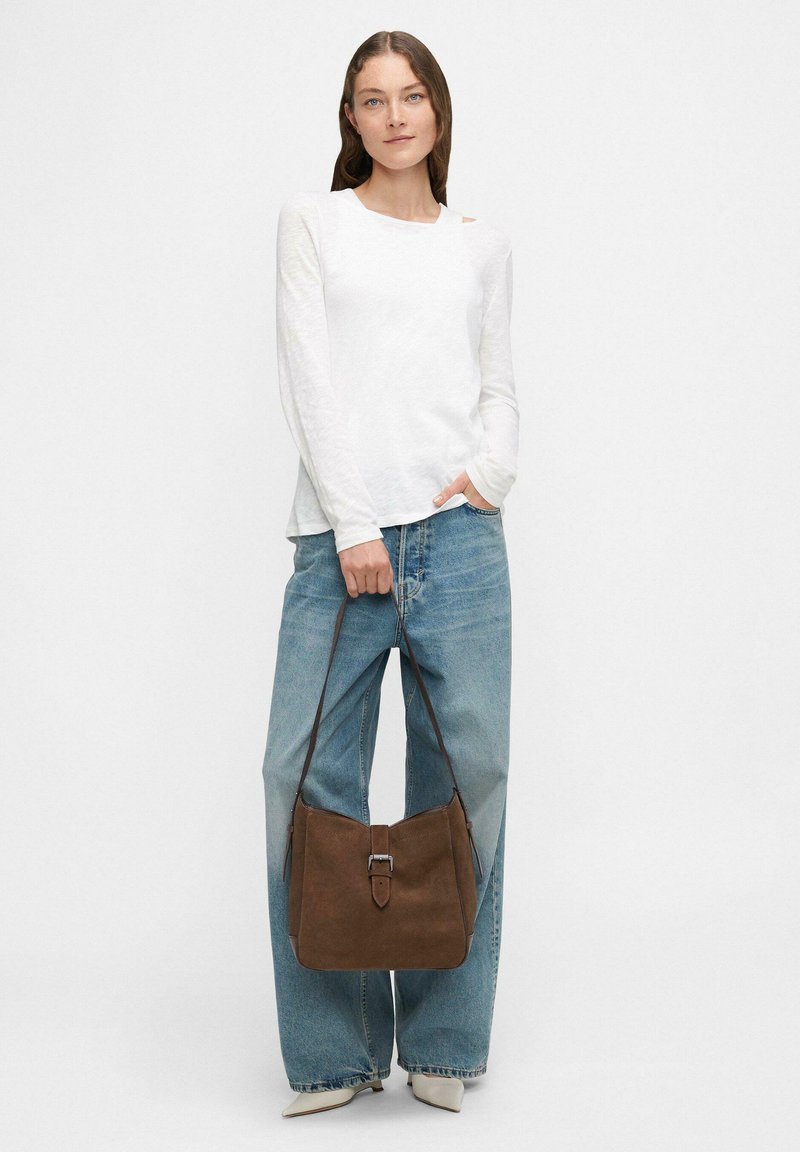 Bolso de ante marrón con detalle de hebilla, combinado con una blusa blanca de manga larga y jeans de tiro alto en azul claro. Diseño sencillo y casual.