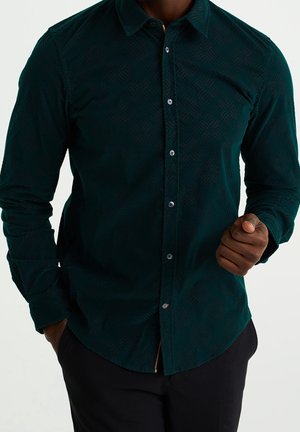 Mørkegrøn skjorte med mønster, lange ærmer, knaplukning foran og en subtil tekstureret design, parret med sorte bukser.