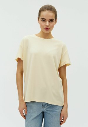 Ung kvinde med samlet hår iført en løs, lysegul kortærmet skjorte og blå jeans mod en ensfarvet hvid baggrund.