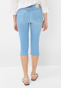Jean capri en denim bleu clair avec une coupe ajustée, longueur mi-mollet, et deux poches arrière, accompagnés de sandales plates.