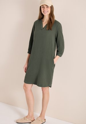 Kobieta w oliwkowozielonej sukience do kolan z rękawami do łokcia, beżowych wsuwanych butach i beżowej czapce z daszkiem, uśmiechająca się z rękami w kieszeniach.