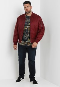 Burgunder bomberjakke med ribbede mansjetter, parret med en svart mønstrete t-skjorte og mørke jeans. Svart sneakers fullfører antrekket.