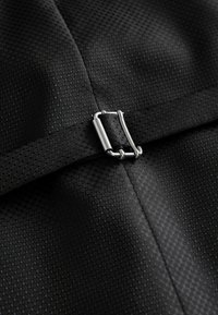 Țesătură neagră texturată cu un model hexagonal. Prezintă o cataramă din metal argintiu atașată la o curea neagră.