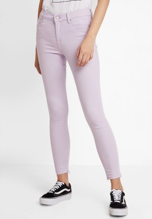 Mujer con vaqueros pitillo de color lila claro con bajos deshilachados y zapatillas negras y blancas, de pie frente a un fondo blanco.