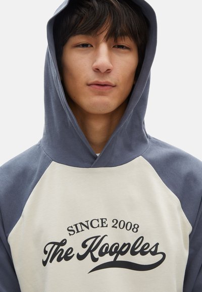 Giovane che indossa una felpa blu e bianca con la scritta "Dal 2008 The Kooples" stampata in nero sul davanti, che guarda in avanti con un'espressione neutra.