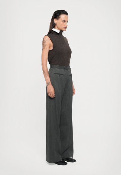 Brown sleeveless turtleneck sweater paired with high-waisted, wide-leg gray trousers featuring a subtle pinstripe, worn with black shoes.