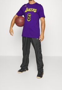 Fialové sportovní tričko s nápisem "LAKERS" a číslem "6" v žluté barvě, doplněné černými, lesklými sportovními kalhotami a černými teniskami, které drží basketbal.