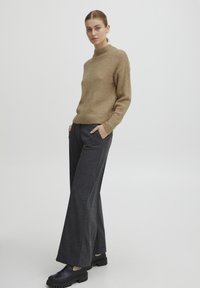 Pull en tricot marron avec col montant, associé à un pantalon large gris foncé et des chaussures noires à enfiler avec des semelles texturées.