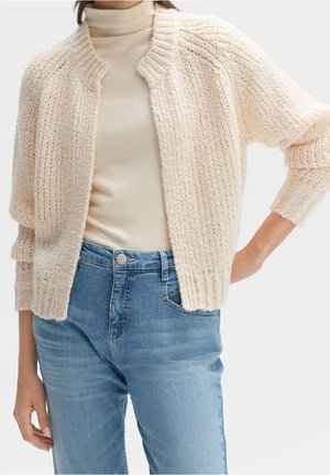 Kobieta ubrana w kremowy, gruby sweter na guziki, nałożony na beżowy golf i jasnoniebieskie, wysokiej talii jeansy, stoi z ręką na biodrze na tle białego tła.