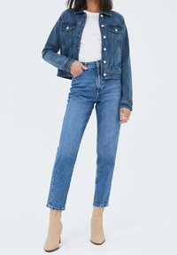 Veste en denim bleu foncé avec un devant à boutons et deux poches poitrine, associée à un jean bleu taille haute et des bottines beige.