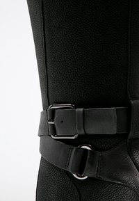 Botte en cuir noir avec finition texturée, dotée d'une large sangle avec une boucle en argent et d'une forme arrondie. Des accents lisses ajoutent des détails.