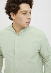 Camisa verde de botones con cuello mao, hecha de tela ligera, que presenta una textura suave y un brillo sutil.