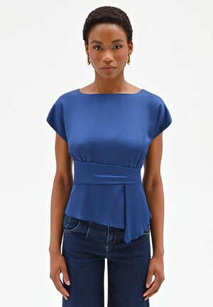 Donna con capelli corti e ricci, che indossa orecchini a cerchio dorati, una blusa asimmetrica blu e jeans blu scuro su uno sfondo bianco.