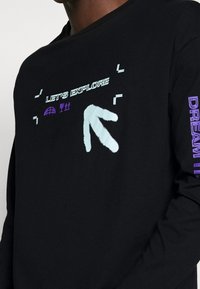 Svart långärmad skjorta med grafisk design som har texten "LÅT OSS UTFORSKA", pilmotiv och lila detaljer på ärmen. Mjuk tyg.